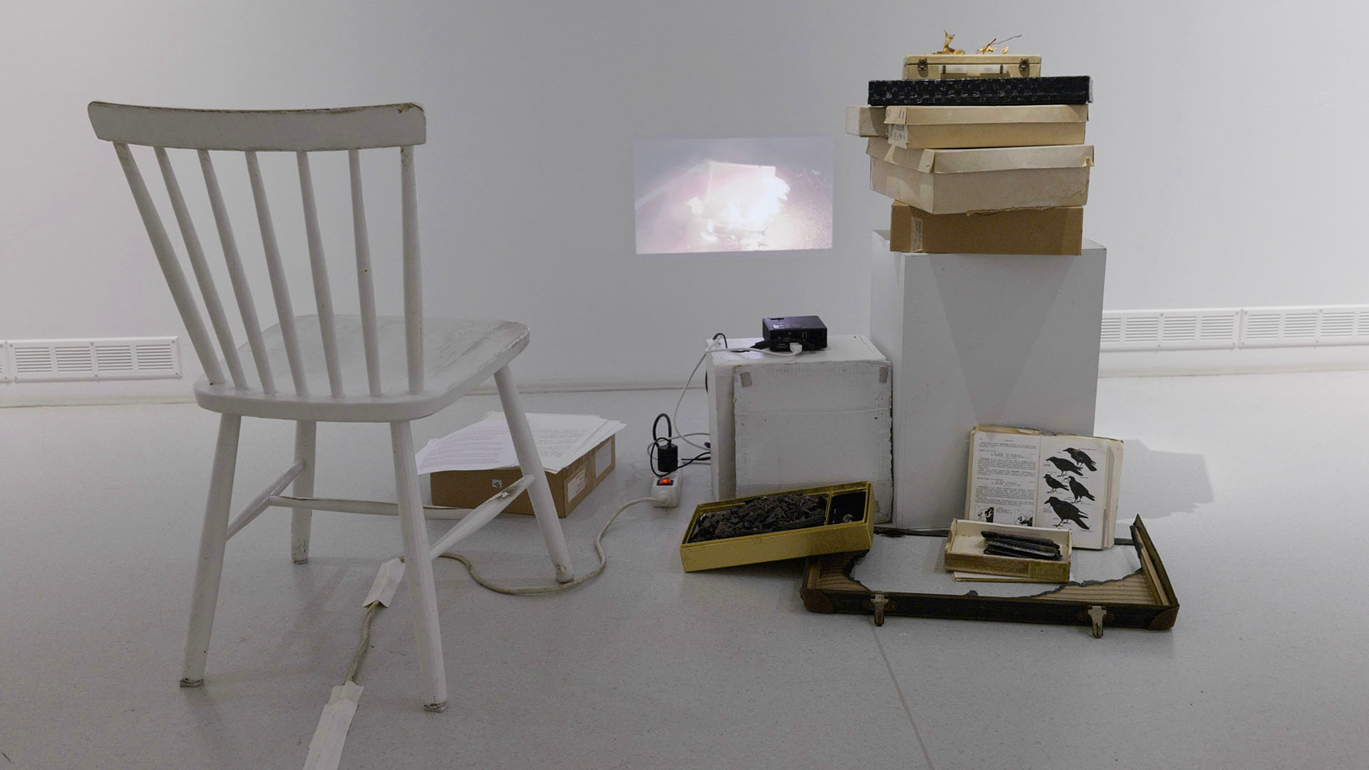 Valkoinen näyttelytila, jossa tuoli, laatikkopino ja pieni videoheijastus seinällä. / A white gallery space with a chair, a stack of boxes and a small video projected on the wall.
